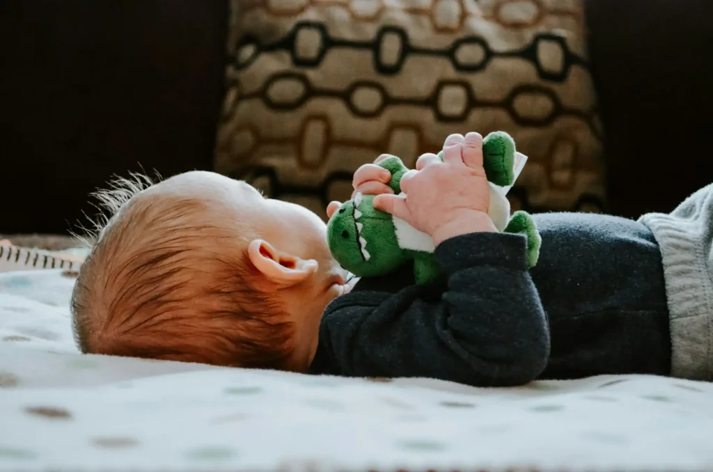 The image shows a close-up of a baby lying on a bed and holding a stuffed dinosaur in his hands.