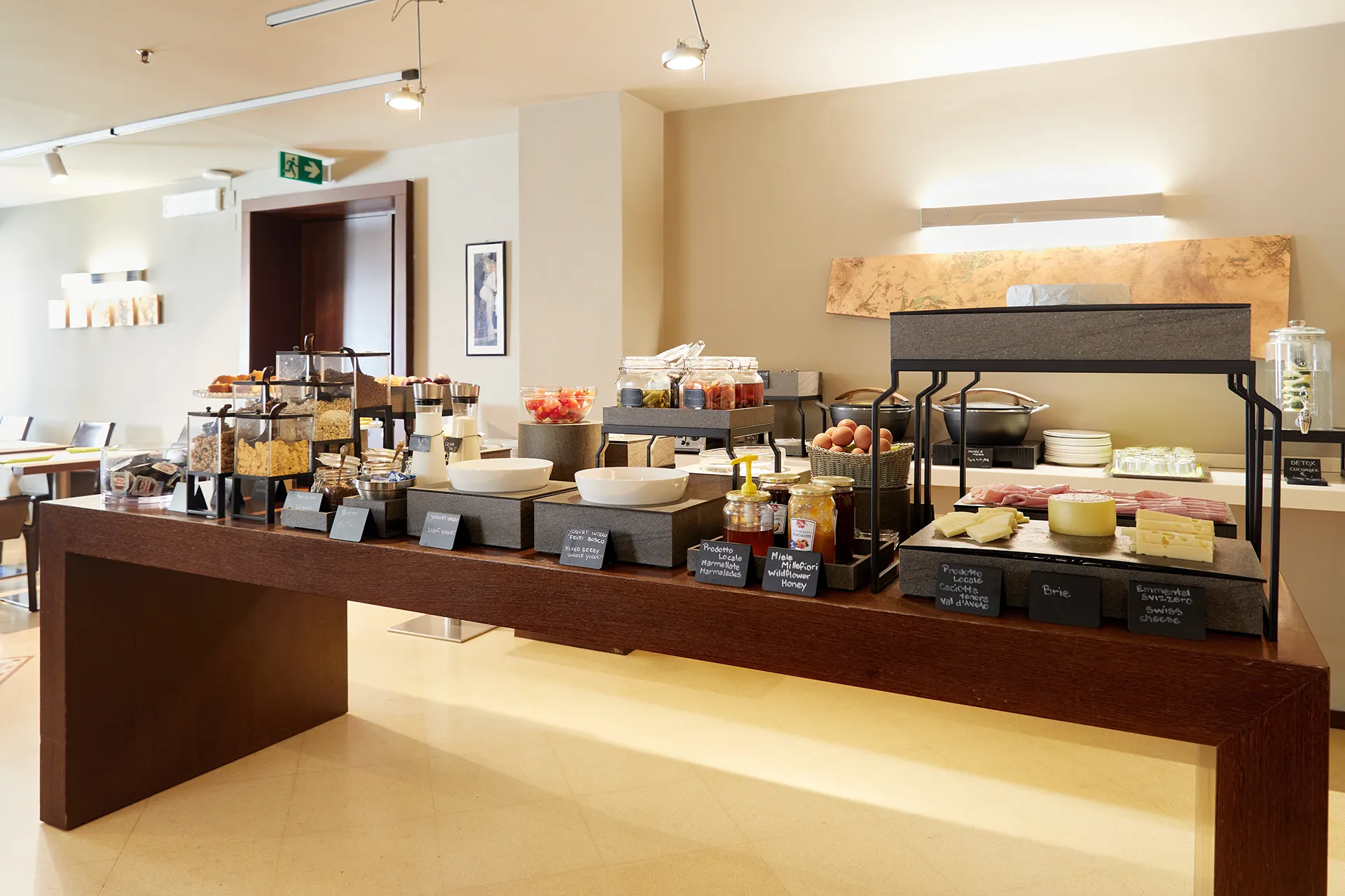The image shows the breakfast buffet at the Hotel Metropoli: on the right, we can see the savory section, with cheeses, cured meats and hard-boiled eggs. On the left, jars of jam and honey, large bowls of yogurt, and, at the back, containers with various cereals.