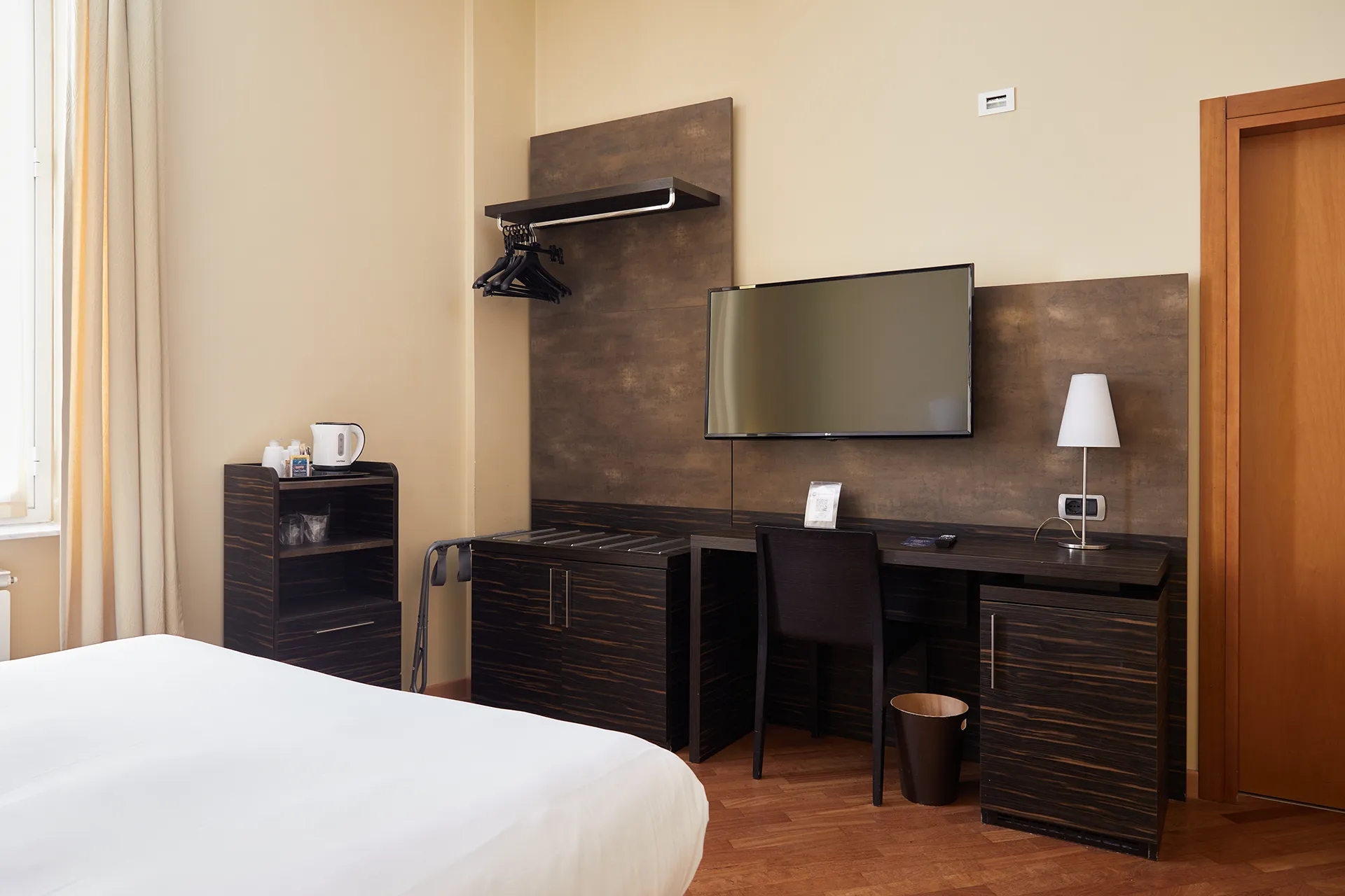 Nell'immagine, un dettaglio della camera Superior dell'Hotel Metropoli: si vede parte del letto e, di fronte, un scrivania ci una sedia e il minibar sotto la scrivania. Alla parete è appesa la TV, mentre di fianco c'è un guardaroba a vista.